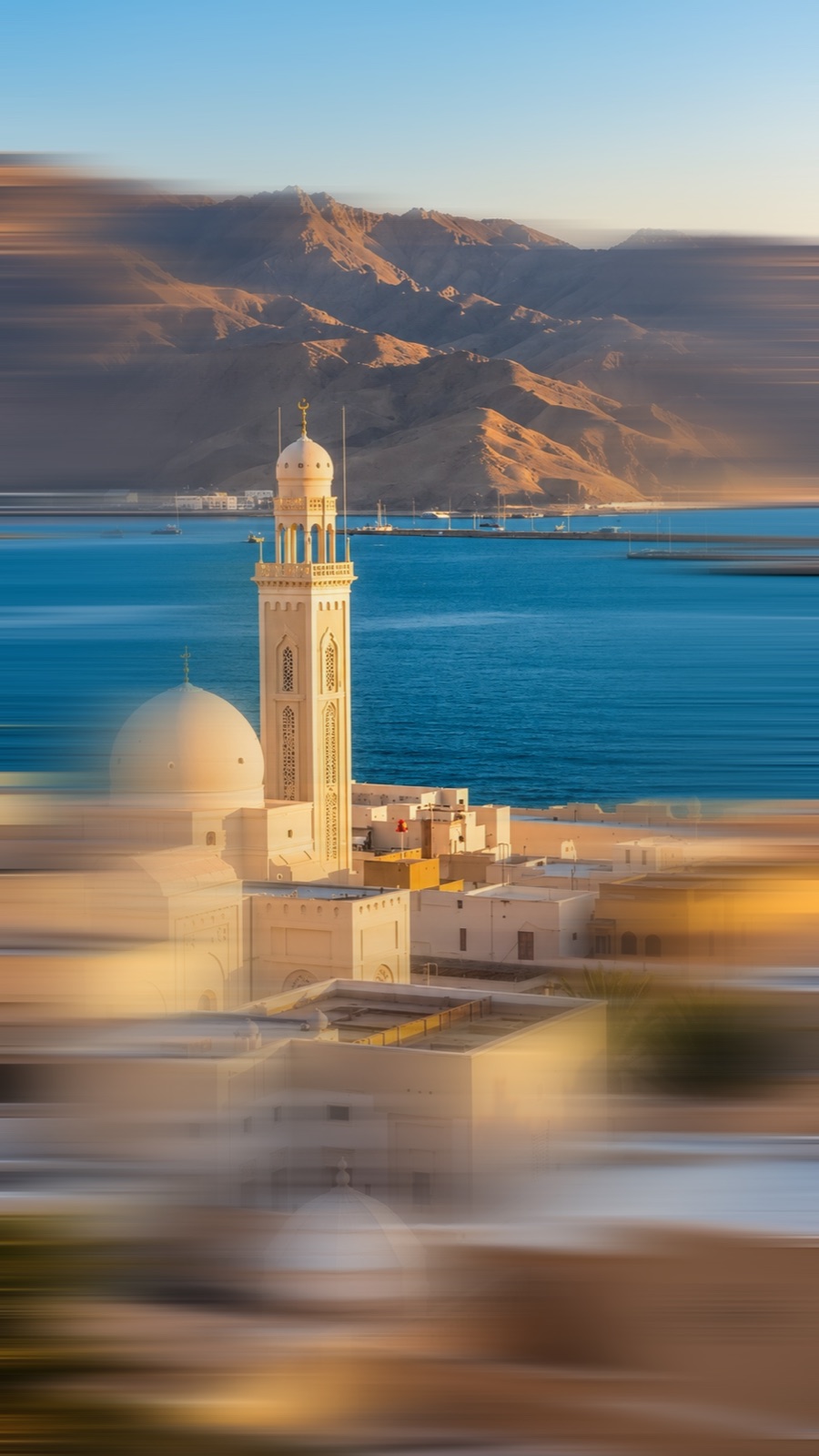 A mosque and minaret by the sea in Muscat, with mountains behind, motion blurred